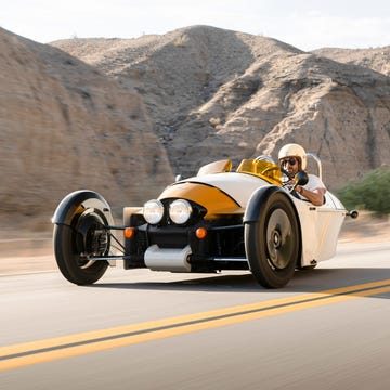 A person driving a yellow 2024 morgan super 3 on a road with mountains in the background